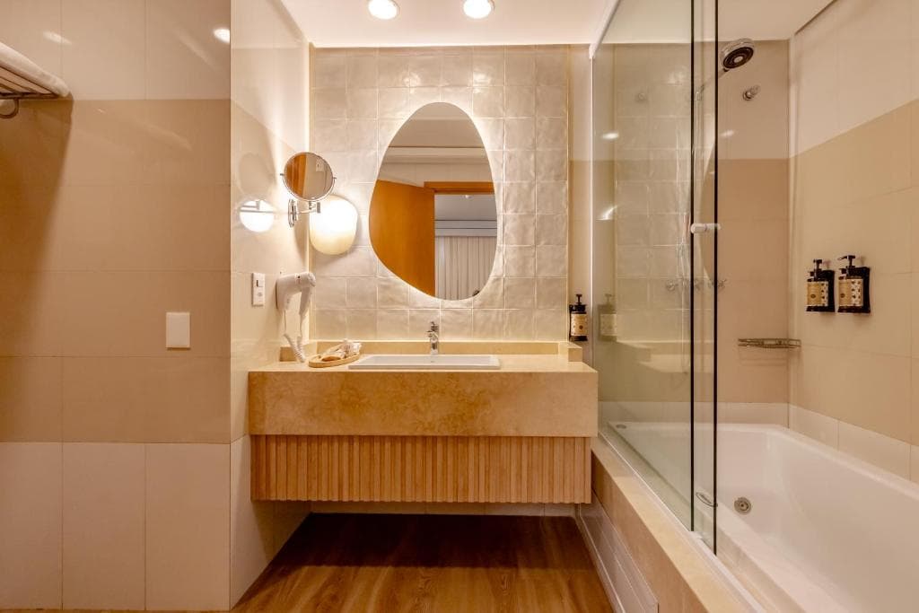 Banheiro moderno com espelho oval iluminado, bancada de mármore e box de vidro transparente.