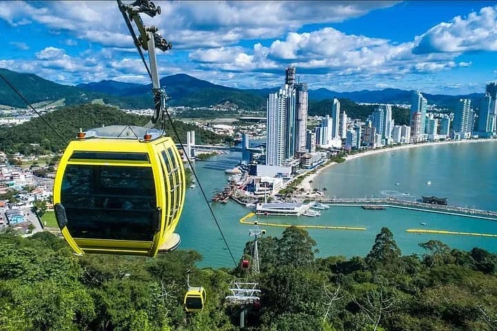 Bondinho amarelo sobrevoando mata verde com vista para arranha-céus e orla de Balneário Camboriú.