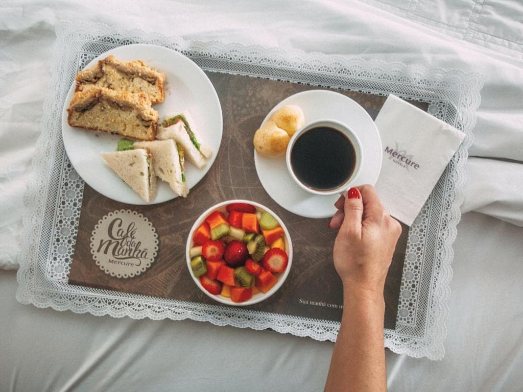 Bandeja de café da manhã com frutas, sanduíches, bolo e café preto sobre lençóis brancos.