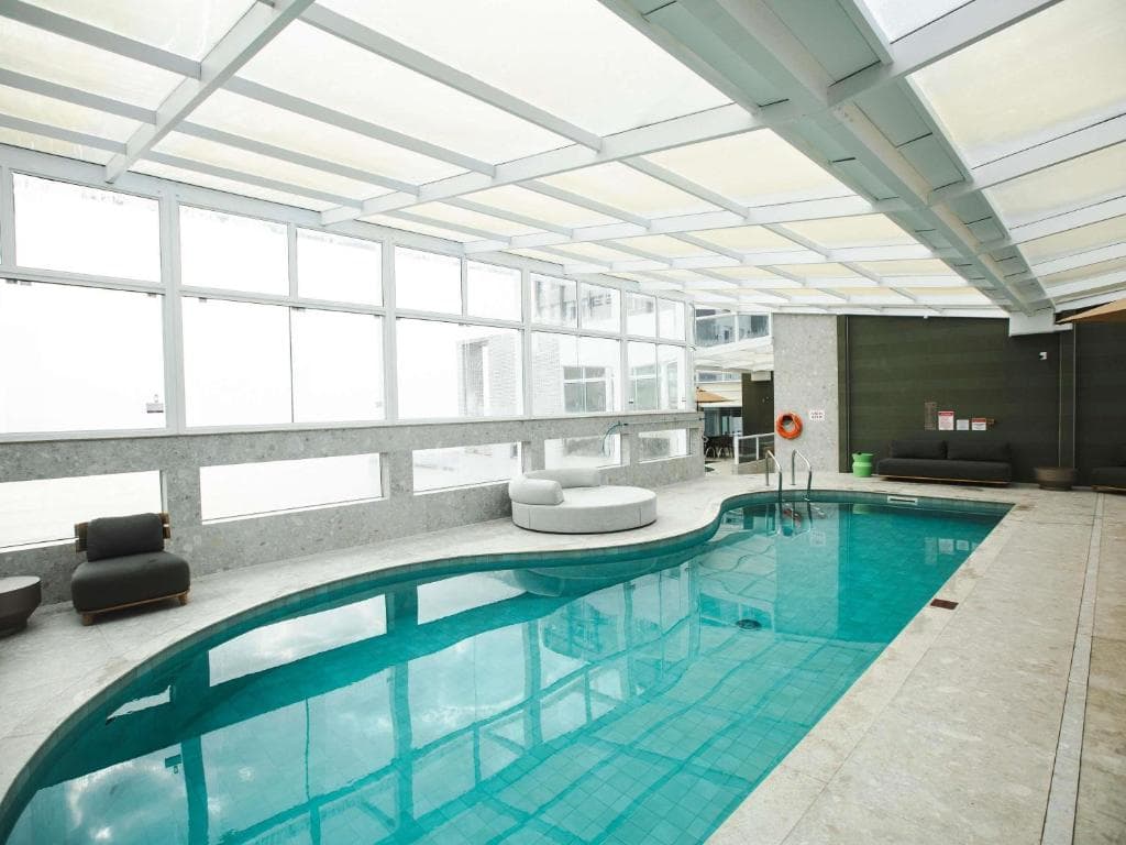 Piscina interna com água azul-turquesa sob teto de vidro e móveis de descanso modernos.