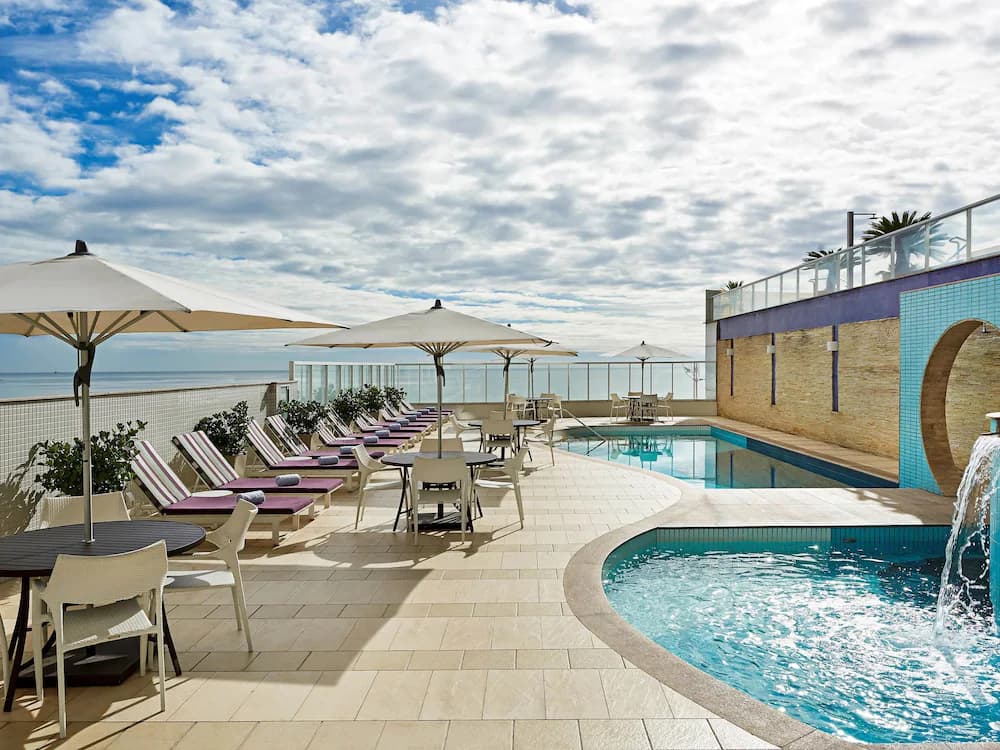 Terraço com piscina, guarda-sóis brancos e espreguiçadeiras roxas com vista para o oceano.
