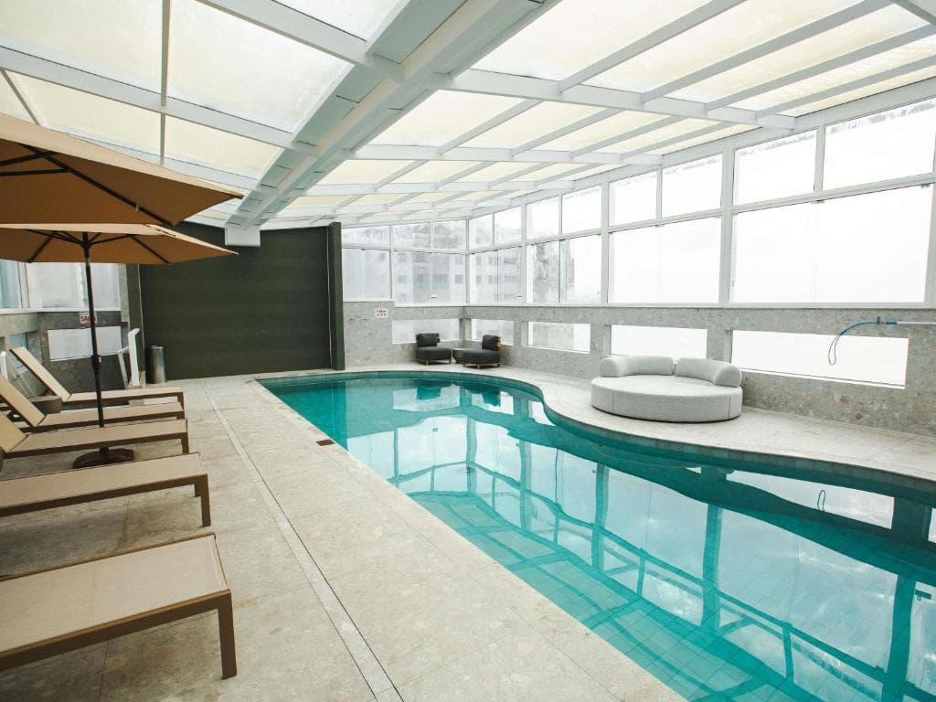 Piscina coberta sob teto de vidro com espreguiçadeiras, guarda-sóis e janelas grandes ao redor.