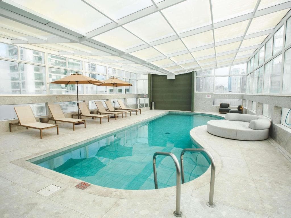 Piscina coberta sob teto de vidro com espreguiçadeiras, guarda-sóis e sofá circular em ambiente moderno.