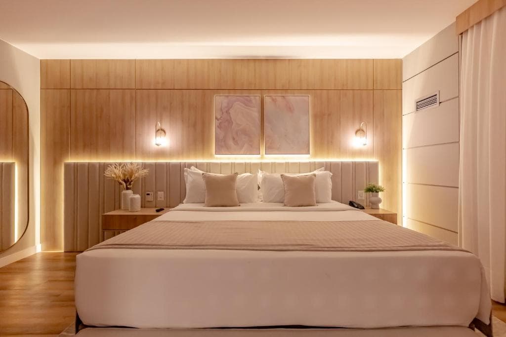 Quarto moderno com cama branca, painel de madeira iluminado e decoração elegante em tons neutros.
