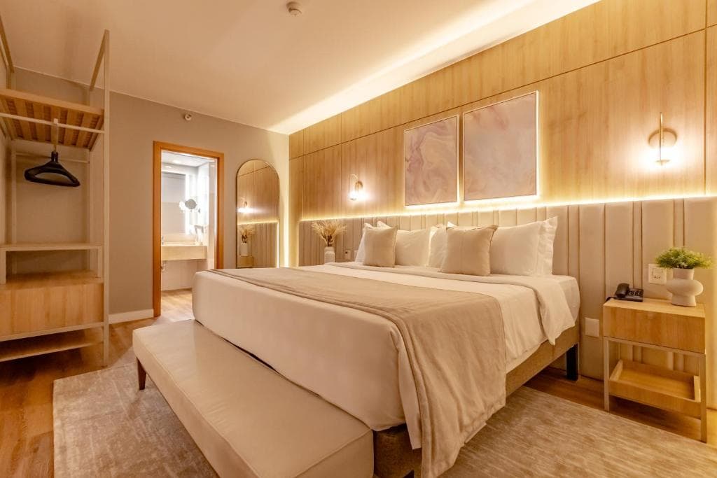 Quarto moderno com cama branca, painel de madeira iluminado, quadros abstratos e tons neutros.