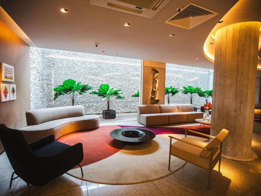 Lounge elegante com sofás curvos, poltronas, tapete circular colorido e plantas verdes em ambiente iluminado.