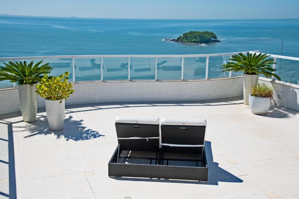 Terraço com espreguiçadeira de vime, vasos de plantas e vista para o mar e ilha.